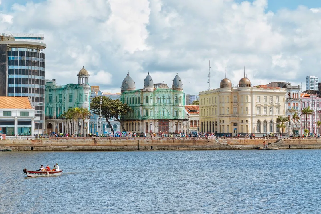 The colorful colonial waterfront of Recife Antigo, with pastel-hued historic buildings reflected on the water and a small boat in the foreground.